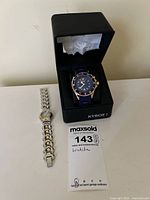 Photo showing Kyboe! watch in black presentation box and Swatch Irony watch with two-tone metal bracelet on table, accompanied by maxsold tag.