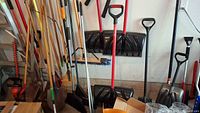 Multiple yard tools in storage against a wall, including shovels, rakes, brooms, and a hedge trimmer all showing signs of soil and use.