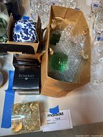 Photo showing a blue and white ceramic decorative ball with floral and bird blue design, packaged in a black Bombay box, alongside a paper bag containing green glass decorative balls, bubble wrapped inside.