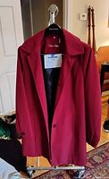 Front view of a red Calvin Klein women's raincoat on a hanger with a black interior lining, showing lapel collar and two front buttons, hanging in a room.