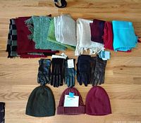 Full spread of 11 scarves in various colors folded and laid out, 5 pairs of women's winter gloves including leather and knit styles, and 3 knit winter hats in different colors, displayed on wooden floor.