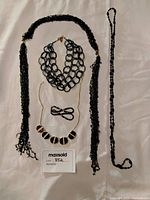 Top-down view of five jewelry pieces on a beige surface, including four necklaces and one bracelet composed of black beads, gold chain, and mixed bead styles.