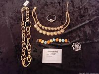 Image of multiple costume jewelry pieces laid out on black background including chain necklace, cluster rhinestone necklace, multi-colored stone bracelet, clear faceted bracelet, and metal floral brooch pin.