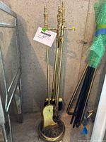 Full view of brass fireplace tools set showing poker, tongs, brush, shovel on a round stand with tripod feet, tagged with lot number