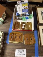 Box with 5 gold single switchplates, 1 gold double plug switchplate, 1 gold triple, 1 double, and 1 brushed gold switchplate on the table with the Kidde alarm in packaging.