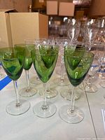 Six green cordial glasses with clear stems and deep green bowls arranged on surface with other glasses in background