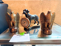 Two bronze-tone metal eagle statues flank a metal dog figurine holding a chain-suspended clock on a glass surface.