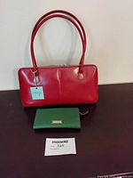 Red leather Hobo handbag with double handles and tag, accompanied by green Cole-Haan wallet on dark surface
