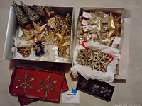 Box with gold glitter star and snowflake ornaments, miniature decorated Christmas trees with bells, small angel figurine, and other assorted gold ornaments.
