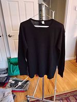 Black cashmere sweater hanging on a metal rack in a room with hardwood flooring and some bags in the background.
