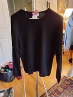 Black crew neck cashmere sweater hanging on a hanger with a pink tag, indoors on wooden floor