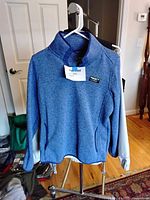 Blue LL Bean pullover sweater hanging on a white hanger against a beige door and chair, showing quarter-zip and tag indicating size large.