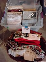 Plastic bin filled with folded linens including more napkins and tablecloths, and a basket with additional folded linen items in various colors and patterns.