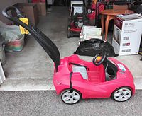 Red plastic car stroller with a black push handle in a garage, showing the side profile of the stroller.