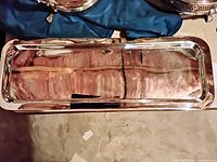 Rectangular polished metal tray with reflective silver-tone finish shown on floor with fabric beneath