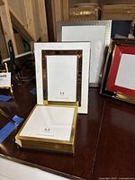 Four photo frames arranged on a table, showing various sizes including 5x5 gold display box frame, 4x6 white standing frame with gold border, 5x7 silver frame with red inner frame, and large 8x10 silver textured frame.