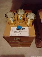 Three gold votive candle holders arranged on top of three stacked boxes labeled as part of the lot set of 63.