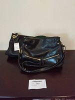 Two black leather purses placed on a surface, one in front showing two front zippered pockets with gold zippers and studded accents, second purse behind with visible tag.