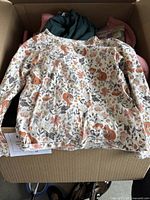 Box containing various toddler girl clothing items including a cream long-sleeve shirt with squirrel print