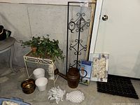 Photo showing all items in the lot arranged on the floor against a gray wall: metal trellis with faux ivy, small metal table, metal watering can, wind chimes in packaging, shell lights, pineapple statue, planters, and yard statue.