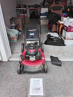 Front view of red Troy-Bilt push mower with Honda engine, owner's manual on ground in front, mower accessories on right side.