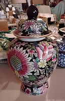 Side view of the floral and butterfly decorated porcelain vase with lid showing colorful pink, white, yellow, and green flowers on a black background.