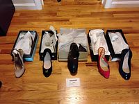Five pairs of women’s shoes lined up outside their boxes on wooden floor. From left to right: beige patent leather heels, black sueded flats, black booties, red sueded slingbacks, and black patent leather heels.