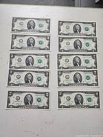 Photo of 10 US $2 bills laid out in three rows showing front side with George Washington's portrait on each bill, confirming currency type, serial numbers visible to show sequential nature.