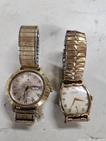 Two gold plated wristwatches, one Stellaris and one Gruen, both with stretch metal bands on a white background.