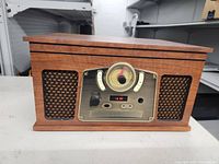 Front view of the wooden cabinet music player showing the central circular dial, control knobs, small digital display, and woven speaker grilles on each side.