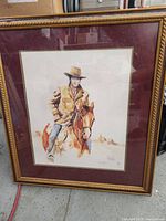Framed watercolor painting of a cowboy on horseback with gold frame and maroon mat