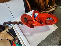 Full side view of the Black & Decker electric hedge trimmer showing the red plastic handle, blade guard, and metal trimming blades on a white surface.