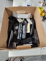 Cardboard box filled with multiple remote controls of different styles and colors, some still in plastic.