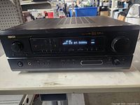 Front view of black Denon AV surround receiver powered on with digital display showing radio frequency and settings.
