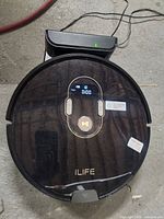 Robot vacuum cleaner with digital display on, showing day, battery, and time, placed on charging dock with power cables visible