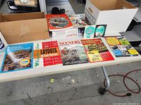 Wide view of table with various small model train related booklets laid out, and model train boxes to the side and behind.