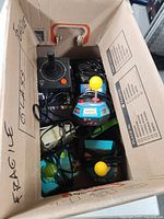 Box containing multiple plug and play game controllers with cables, including a black joystick and colorful controllers with yellow ball joysticks, all untested.