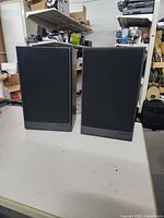 Two Polk Audio speakers side by side with grilles on, showing their rectangular shape and black finish on a white table with cluttered background.