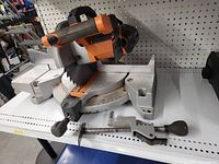Full view of Rigid 10-inch angle chop saw with clamp and adjustable base for angle cuts, showing saw blade and handle, sitting on white perforated shelf.