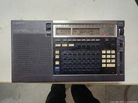 Front view of Sony ICF-2010 radio showing speaker grill, digital clock display, frequency readout, and numeric keypad with tuning buttons.