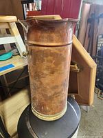 Tall copper umbrella stand with tarnish and patina, placed on a stool with some surrounding wooden furniture.