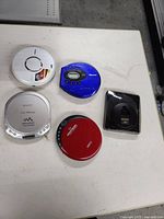 Five portable CD players arranged on a white surface, each with visible brand and buttons for operation.