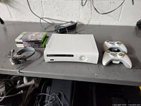 Full setup showing Xbox 360 console in white, stack of 4 game cases, wired headset, and two wireless controllers on a gray table.