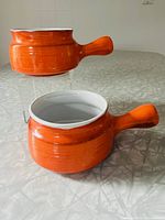 Pair of orange drip lug-handled soup bowls shown together on a table, highlighting their shape and color.