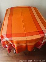 Full tablecloth draped over small table showing vibrant shades of orange with broad and narrow stripes in red, beige, and orange tones.