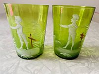 Close-up photo showing two green glass tumblers with hand-painted white enamel figures depicting children in landscape scenes. The tumblers have a bright translucent green color and detailed Mary Gregory style artwork.