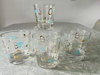 Four clear glasses with mid-century Atomic Amoeba (Boomerang) pattern in gold and turquoise. Arranged with three at front and one elevated behind.