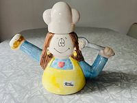 Front view of the hand-painted ceramic teapot showing the female chef character with outstretched arms, holding a donut on the spout hand and an ice cream scoop on the handle hand, wearing yellow apron and white chef hat.