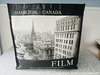 Front view of black vinyl tote bag showing black and white photo of historic Hamilton architecture and printed text 'HAMILTON | CANADA' and 'FILM'.