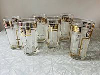 Six vintage Dominion Glassware beverage glasses arranged closely together, showing gold Greek key border and white etched fern designs.